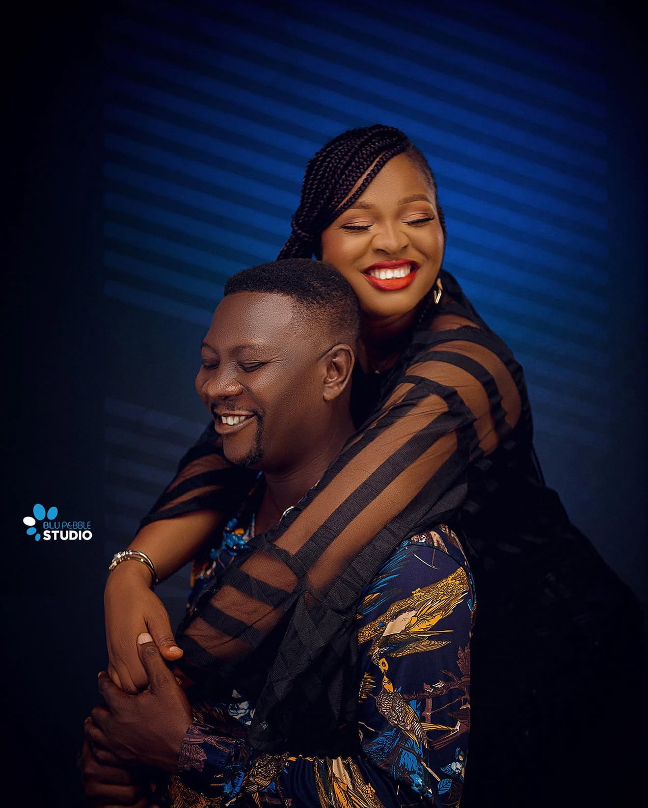 Couple portrait with a woman embracing a man from behind against a deep blue studio backdrop
