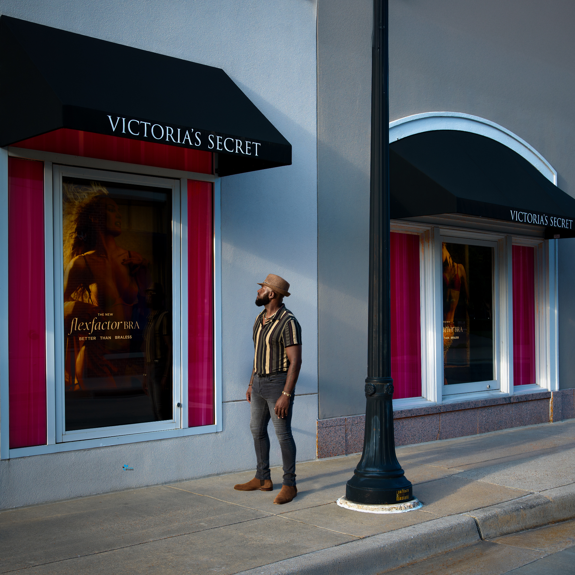 Man in a striped shirt and fedora standing in late-afternoon light outside a Victoria's Secret storefront