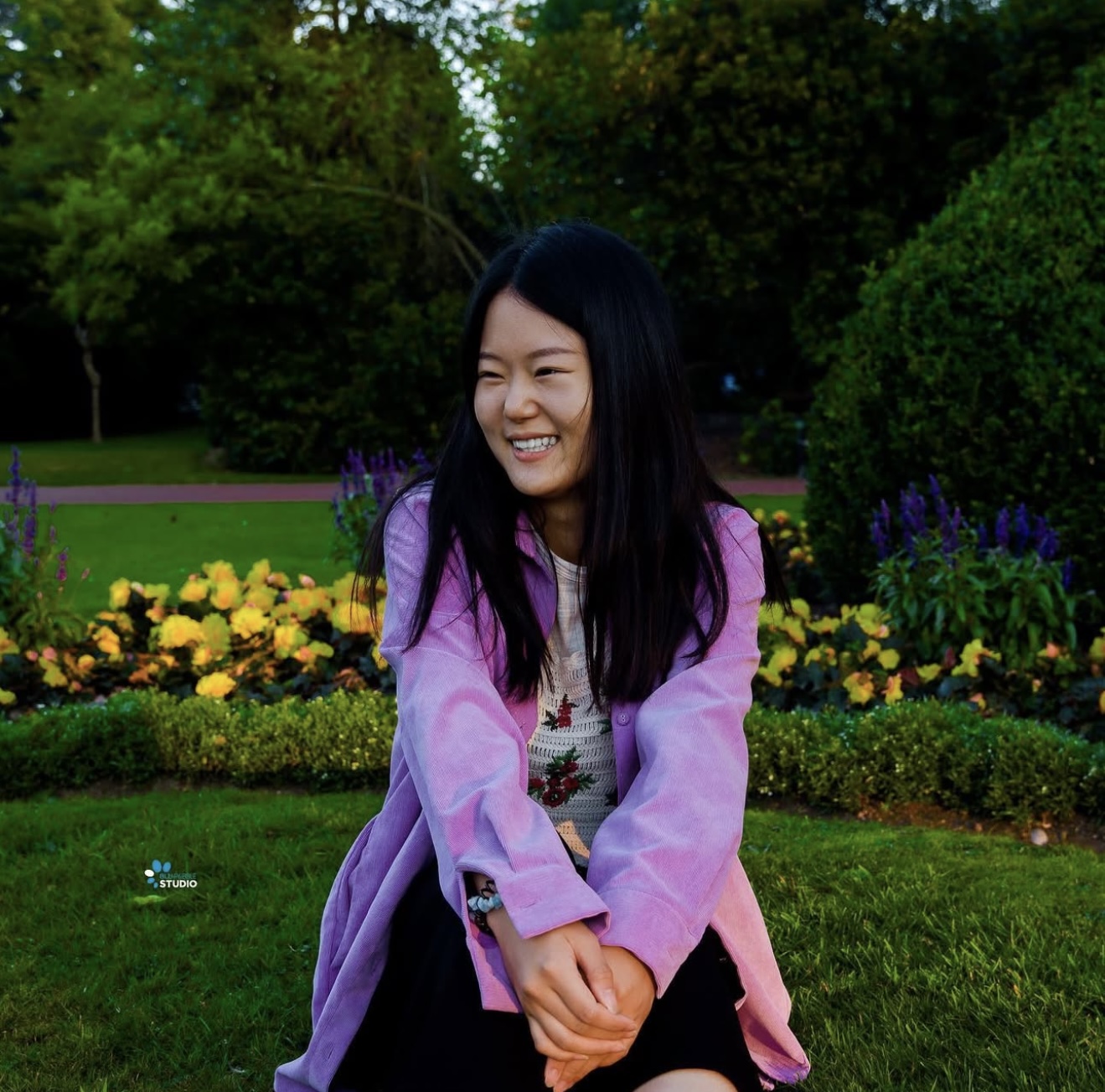 Young woman in a lilac corduroy shirt laughing while seated on grass in front of a flower bed
