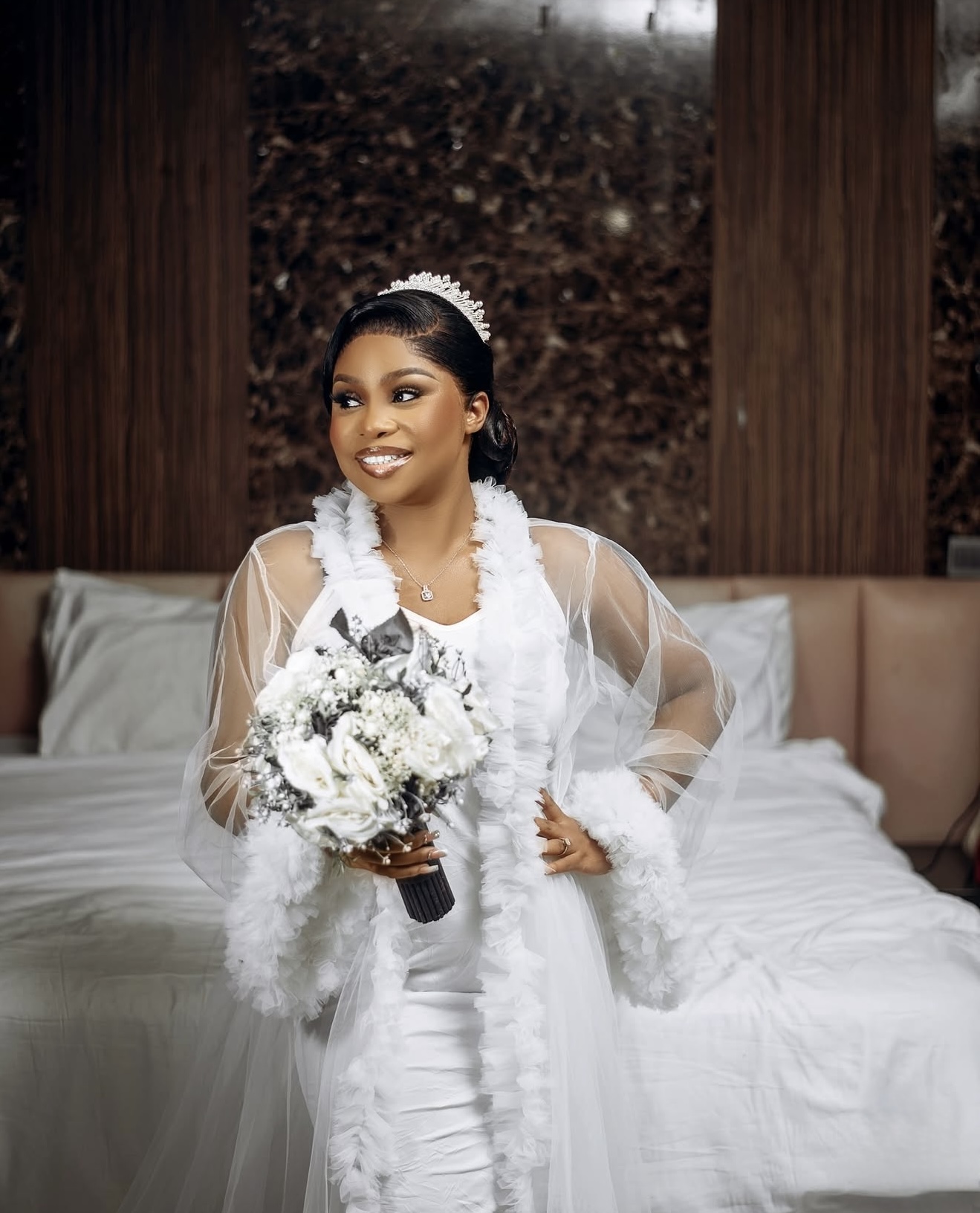 Bride in a white feathered robe holding a bouquet while standing in a bedroom