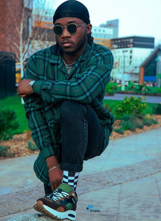 Man in a green plaid shirt, durag and round sunglasses crouching on a city plaza ledge