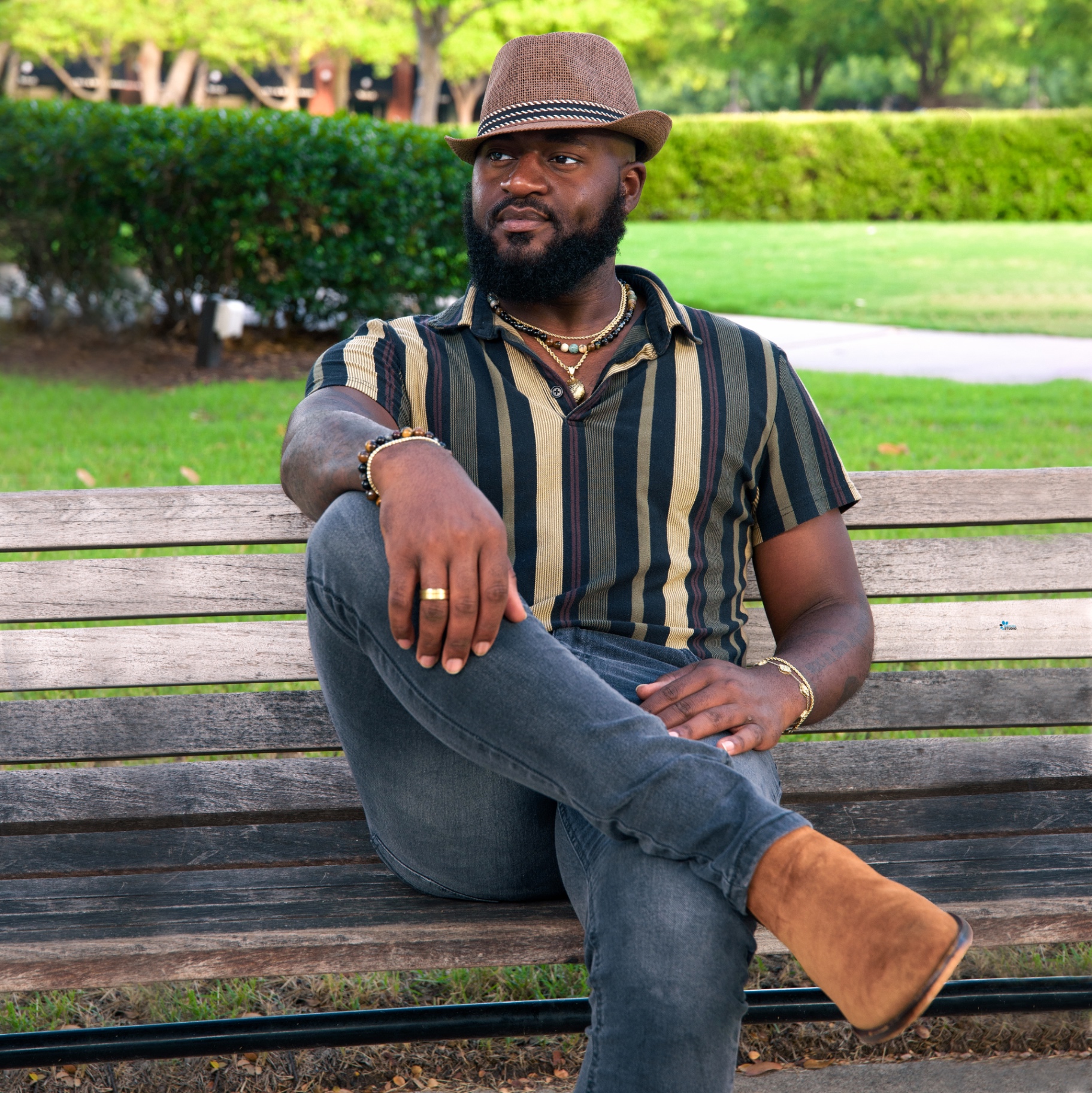 Man in a striped knit polo and fedora seated on a weathered park bench with green hedges behind
