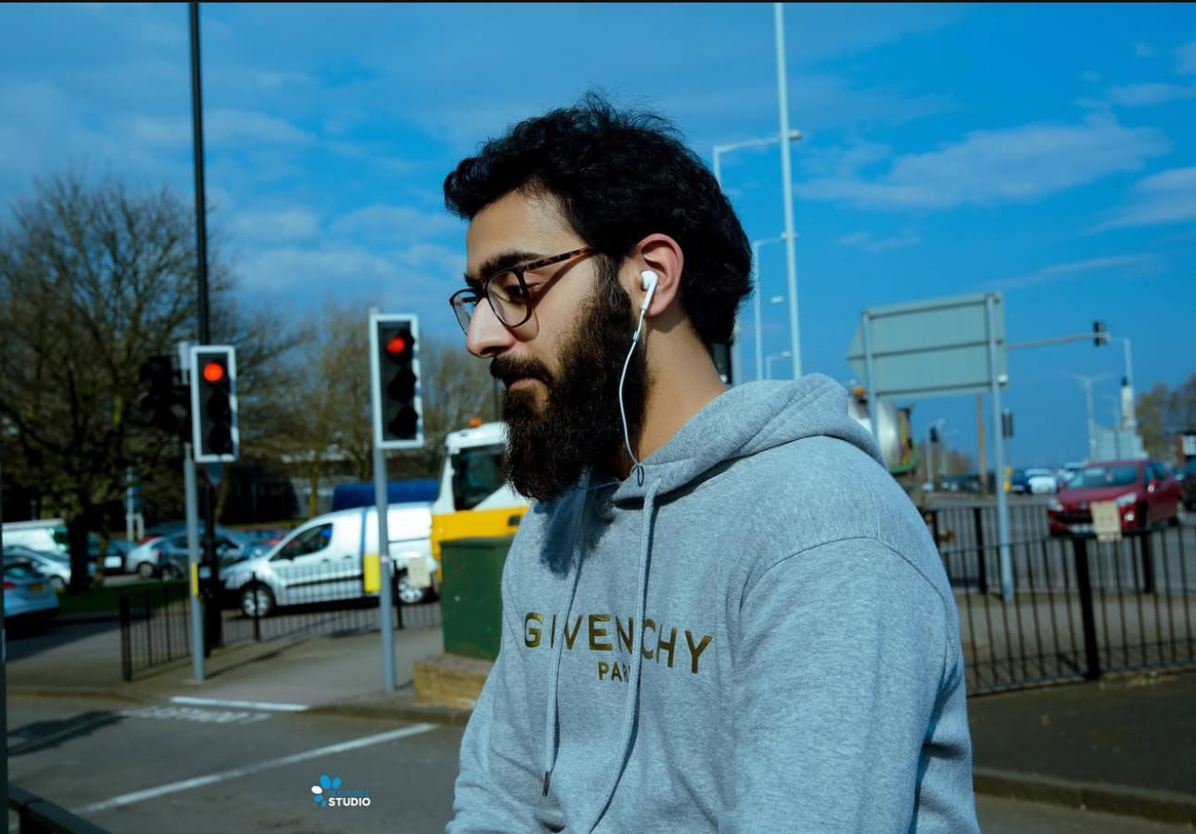 Bearded man in a grey Givenchy hoodie in profile with earphones in, at a sunlit city crossing