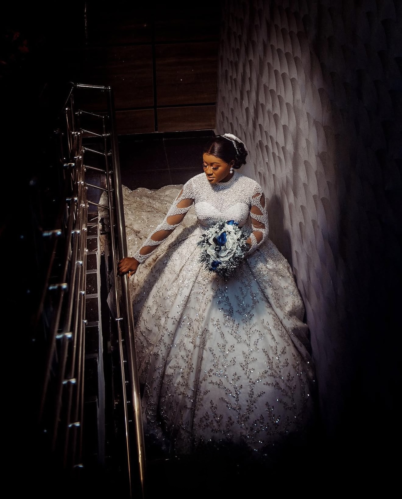 Bride in a beaded wedding gown descending a staircase with a blue and white bouquet