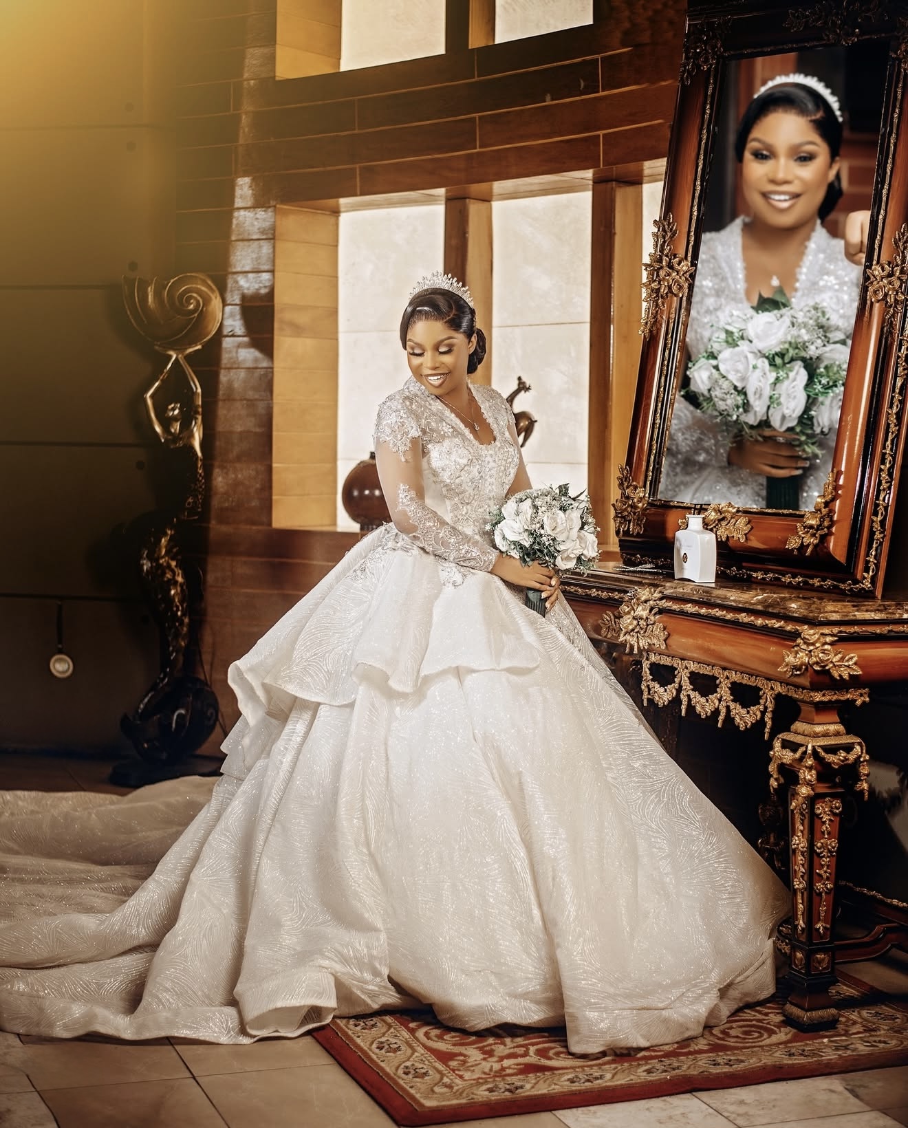 Bride in a full white wedding gown seated by an ornate mirror holding a white bouquet
