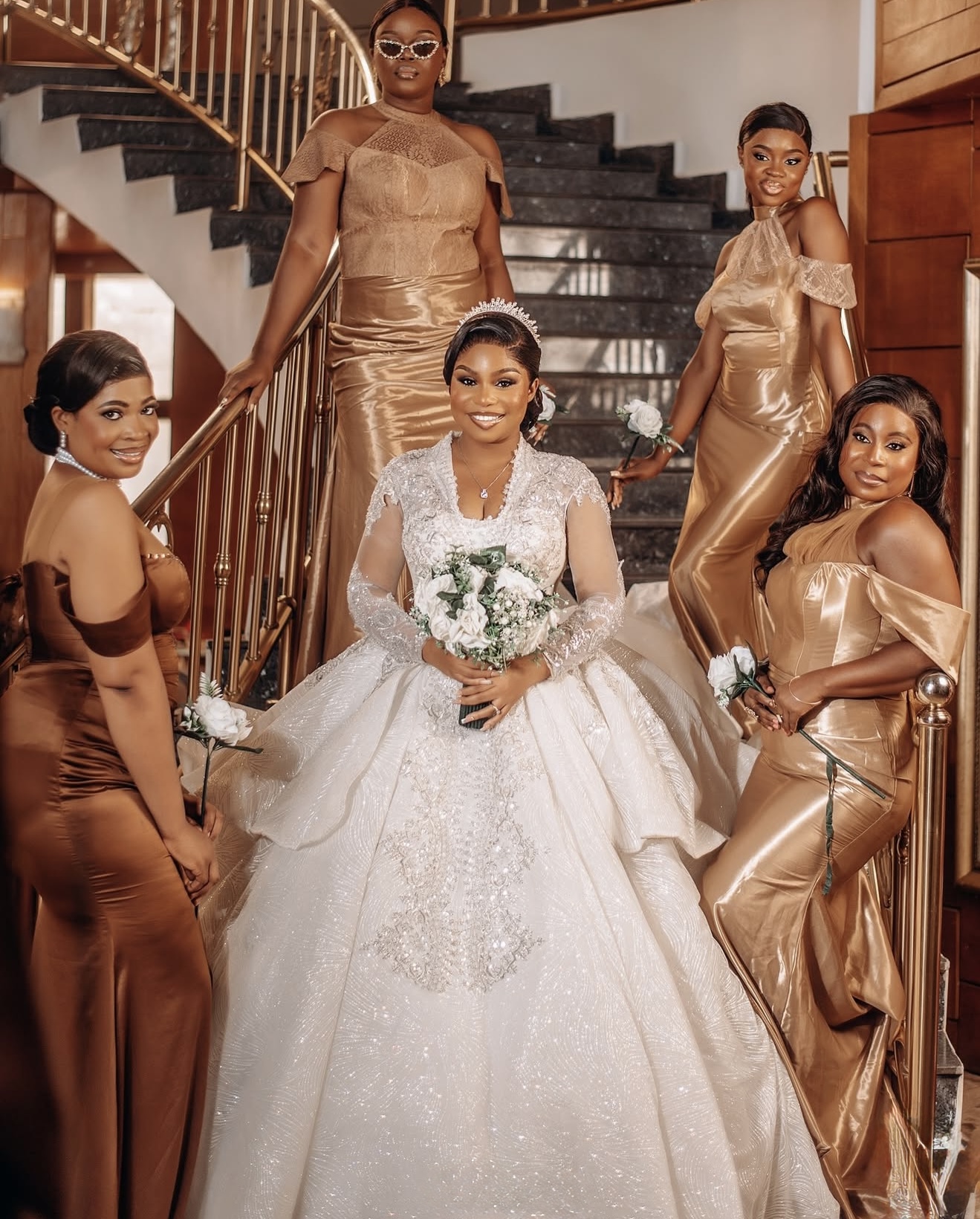 Bride in a white gown surrounded by bridesmaids in gold dresses on a curved staircase
