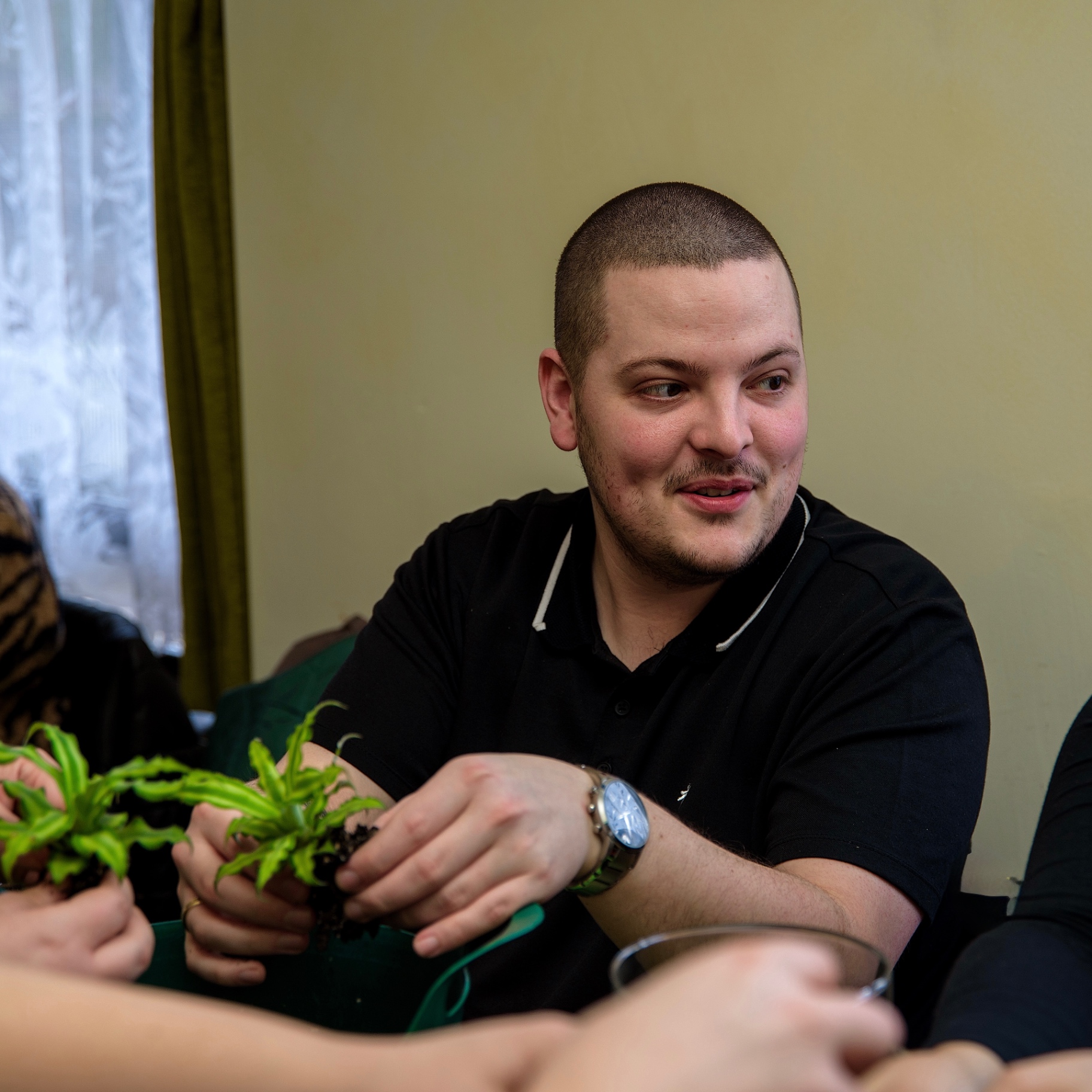 Man in a black polo shirt smiling as he passes a fern seedling across the workshop table