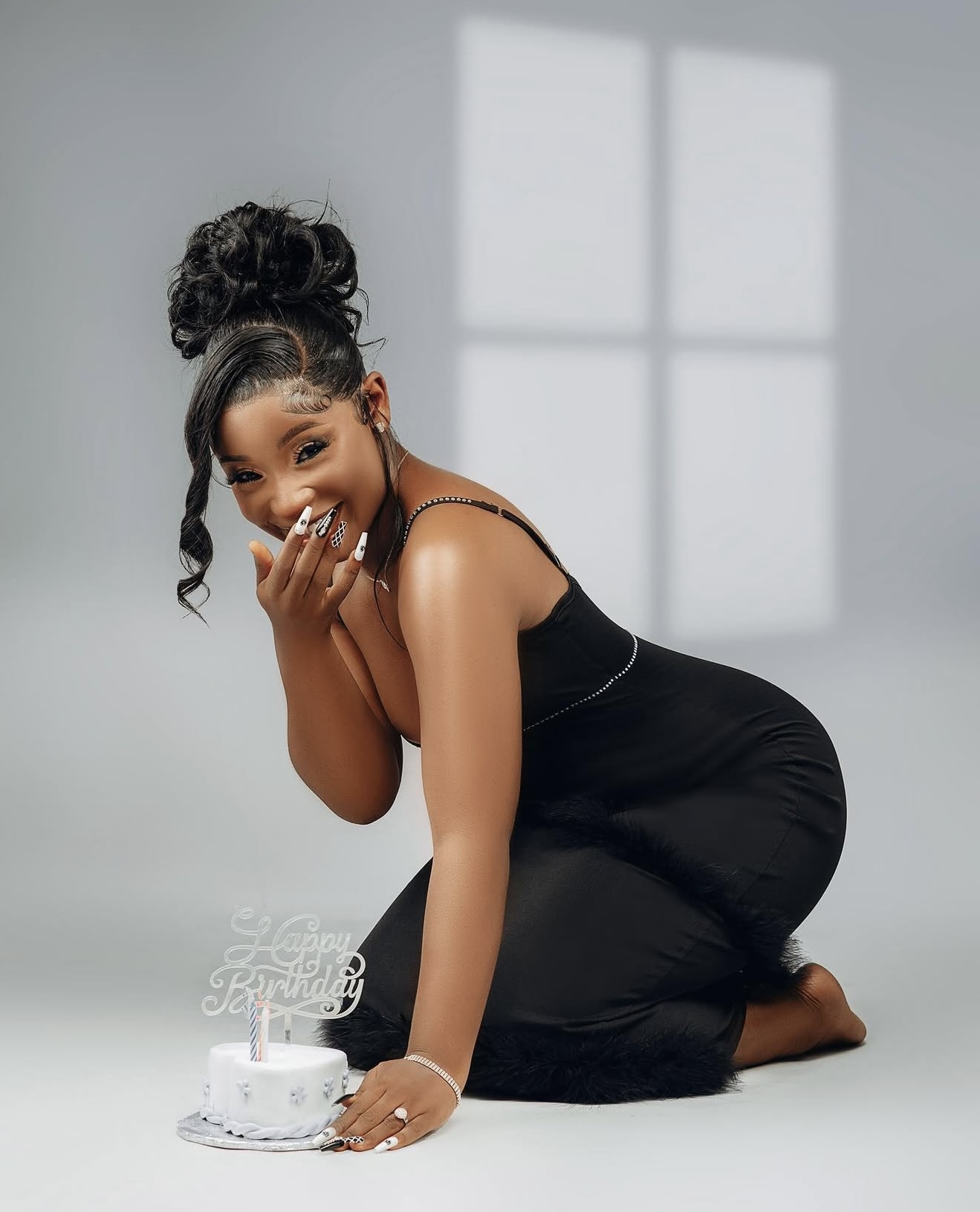 Woman in a black dress kneeling beside a small white birthday cake in a bright studio